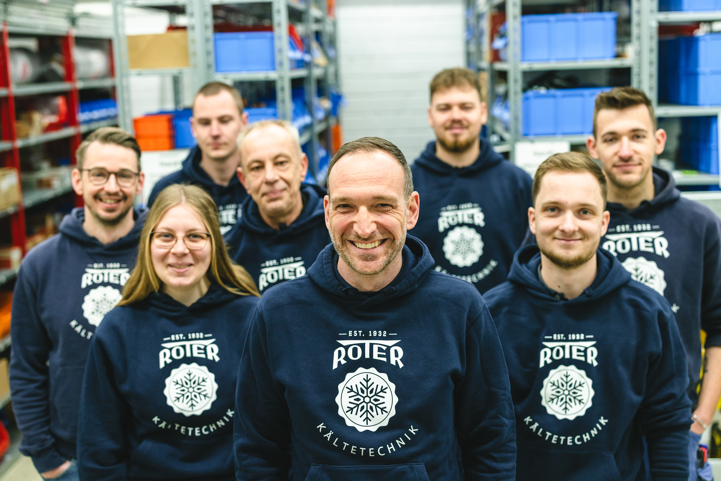 viel liebe x roter kaeltetechnik – B content 11 0515 Team von Roter Kältetechnik in Firmen-Hoodies vor Regalen im Lager – neun Mitarbeitende lächeln in die Kamera und zeigen Teamgeist und moderne Handwerkskultur.