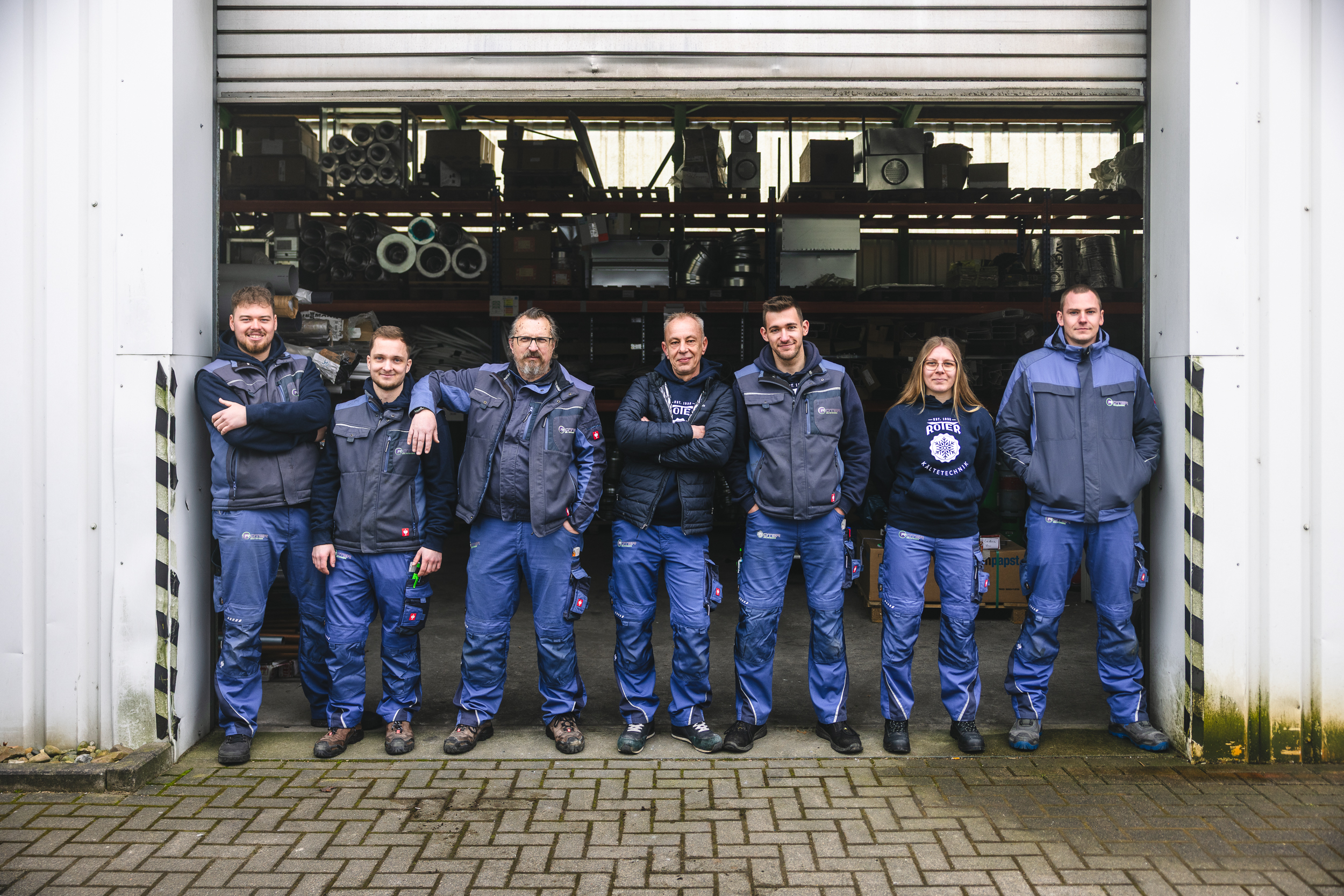 viel liebe x roter kaeltetechnik – B content 18 0658 Team von Roter Kältetechnik vor offener Werkstatthalle – Mitarbeiter:innen in blauer Arbeitskleidung posieren gemeinsam vor Regalen mit Kälte- und Lüftungstechnik.