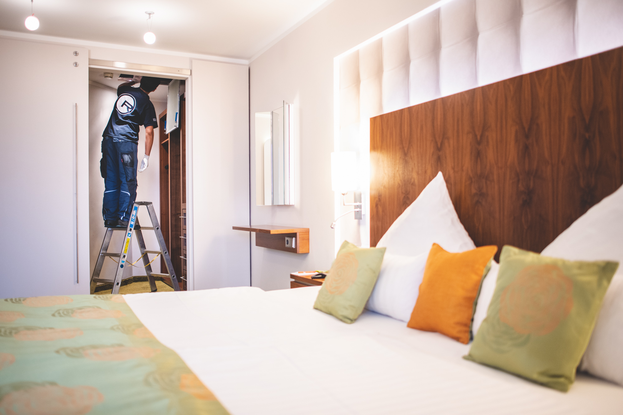 Hotel Zimmer „Handwerker auf Leiter arbeitet im Hotelzimmer, Bett im Vordergrund.“