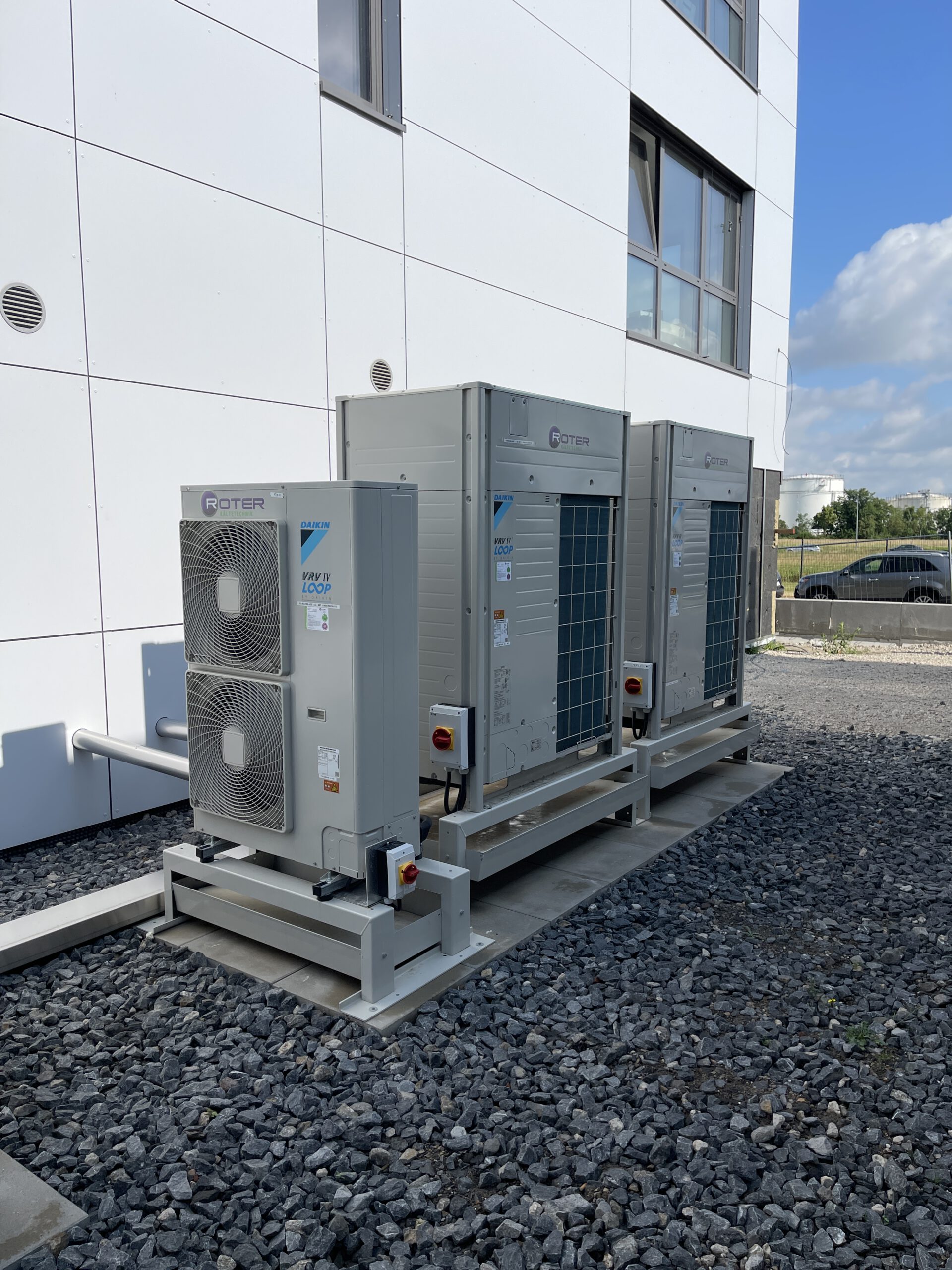 VRV 2 Drei graue Daikin VRV IV Klimaanlagen auf Gestell vor weißem Bürogebäude.